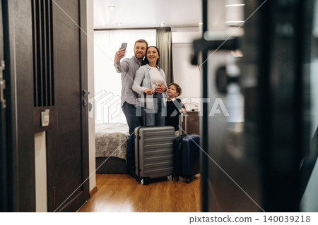 Family of three taking a selfie in a hotel room with luggage, showcasing a modern interior design and bright natural light from large windows Family of three taking a selfie in a hotel room with luggage, showcasing a modern interior design and bright natural light from large windows 140039218