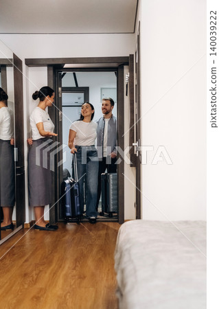 Hotel reception area with a female staff member greeting two guests carrying luggage, modern interior design featuring wooden flooring and minimalist decor 140039222