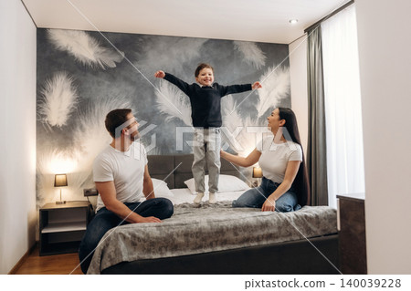 Family enjoying time together in a hotel room with a child jumping on the bed, modern decor featuring feather-patterned wall and soft lighting 140039228