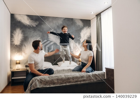 Family enjoying time together in a modern hotel room with a child jumping on the bed, featuring stylish decor and soft lighting in a cozy atmosphere 140039229
