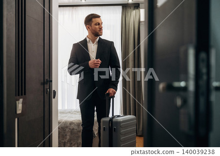 Businessman in formal attire holding a suitcase stands at the entrance of a modern hotel room with a bed and curtains visible in the background Businessman in formal attire holding a suitcase stands at the entrance of a modern hotel room with a bed and curtains visible in the background 140039238