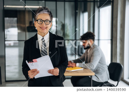 Mature businesswoman in black suit holding documents smiles confidently in modern office with male colleague working at desk in background Mature businesswoman in black suit holding documents smiles confidently in modern office with male colleague working at desk in background 140039590