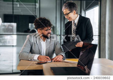 Business professionals engaged in discussion at a modern office workspace, with a laptop and documents on the table, large windows providing natural light 140039602