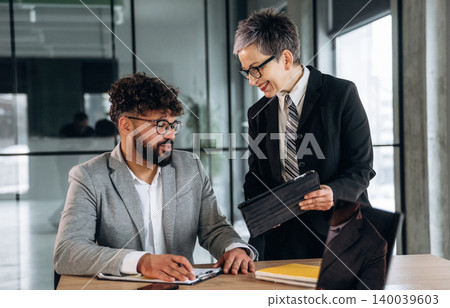 Business meeting in modern office with two professionals discussing project details at a wooden table, digital tablet and documents visible in a bright workspace 140039603