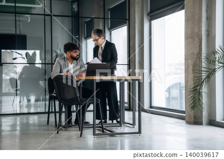 Business meeting in modern office with male and female professionals discussing documents at a table, large windows and greenery visible in the background Business meeting in modern office with male and female professionals discussing documents at a table, large windows and greenery visible in the background 140039617