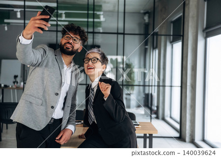 Two business professionals taking a selfie in a modern office setting with large windows and a wooden table, showcasing a collaborative work environment Two business professionals taking a selfie in a modern office setting with large windows and a wooden table, showcasing a collaborative work environment 140039624