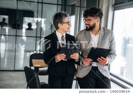 Business professionals in formal attire discussing project details while holding tablets inside a modern office with large windows and a collaborative workspace 140039634