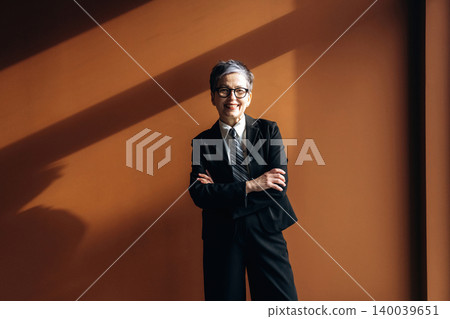 Senior Asian businesswoman in black suit stands confidently with arms crossed against a warm orange wall, showcasing professional attire and modern office environment Senior Asian businesswoman in black suit stands confidently with arms crossed against a warm orange wall, showcasing professional attire and modern office environment 140039651