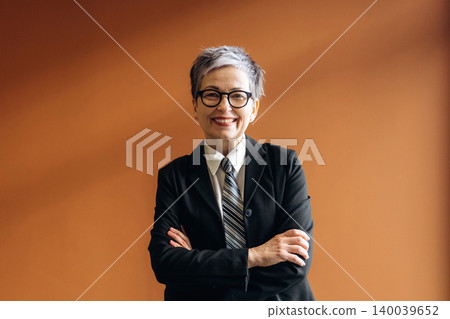 Businesswoman with short gray hair and glasses wearing a black suit and tie, standing confidently with arms crossed against a warm orange wall in a modern office setting Businesswoman with short gray hair and glasses wearing a black suit and tie, standing confidently with arms crossed against a warm orange wall in a modern office setting 140039652