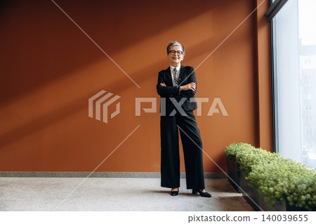 Businesswoman in black suit stands confidently with arms crossed in modern office space featuring a warm brown wall and green plants along the floor Businesswoman in black suit stands confidently with arms crossed in modern office space featuring a warm brown wall and green plants along the floor 140039655