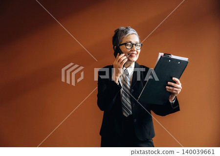 Businesswoman with short gray hair in a black suit holds a clipboard while talking on a smartphone against a warm brown wall in a modern office setting Businesswoman with short gray hair in a black suit holds a clipboard while talking on a smartphone against a warm brown wall in a modern office setting 140039661