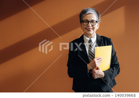 Senior Asian businesswoman in a black suit holding a yellow folder, smiling confidently against a warm orange wall with diagonal light shadows 140039668