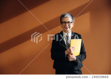 Senior businesswoman with short gray hair wearing a black suit and glasses holds a yellow folder while standing against a brown wall in a modern office setting Senior businesswoman with short gray hair wearing a black suit and glasses holds a yellow folder while standing against a brown wall in a modern office setting 140039669