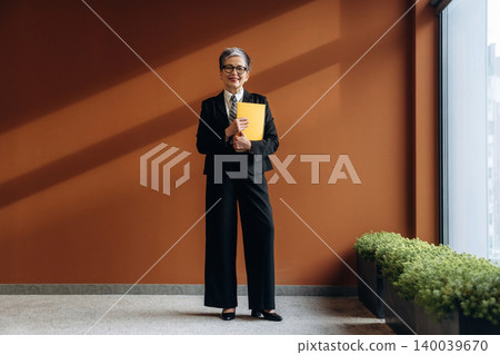 Senior businesswoman in a black suit holding a yellow folder stands confidently in a modern office space with large windows and a brown accent wall Senior businesswoman in a black suit holding a yellow folder stands confidently in a modern office space with large windows and a brown accent wall 140039670