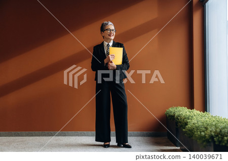 Businesswoman with short gray hair wearing a black suit holds a yellow folder while standing in a modern office with a brown wall and green plants Businesswoman with short gray hair wearing a black suit holds a yellow folder while standing in a modern office with a brown wall and green plants 140039671