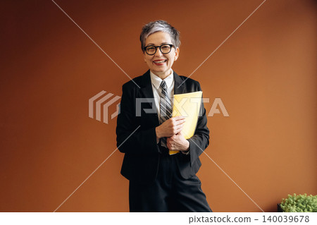 Senior businesswoman with short gray hair wearing a black suit and glasses holds yellow folder while standing in modern office with brown wall and green plant Senior businesswoman with short gray hair wearing a black suit and glasses holds yellow folder while standing in modern office with brown wall and green plant 140039678