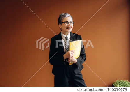 Senior businesswoman in black suit and glasses holding yellow folder, smiling confidently against a warm brown wall in a modern office setting Senior businesswoman in black suit and glasses holding yellow folder, smiling confidently against a warm brown wall in a modern office setting 140039679