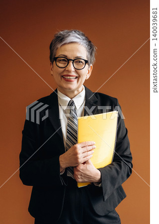 Businesswoman with short gray hair wearing glasses holds a yellow folder while smiling in a professional suit against a brown background in an office setting Businesswoman with short gray hair wearing glasses holds a yellow folder while smiling in a professional suit against a brown background in an office setting 140039681