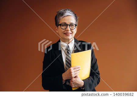 Senior businesswoman with short gray hair wearing glasses and a black suit holds a yellow notebook while standing against a brown background in an office setting Senior businesswoman with short gray hair wearing glasses and a black suit holds a yellow notebook while standing against a brown background in an office setting 140039682