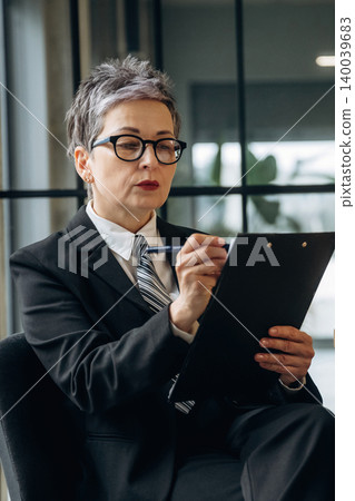 Businesswoman with short gray hair wearing glasses takes notes on a clipboard while seated in a modern office environment with large windows and greenery visible Businesswoman with short gray hair wearing glasses takes notes on a clipboard while seated in a modern office environment with large windows and greenery visible 140039683