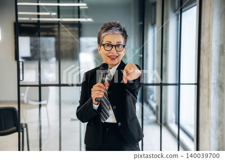Businesswoman in black suit holding microphone gestures while speaking in modern office with glass walls and minimalistic furniture in the background 140039700