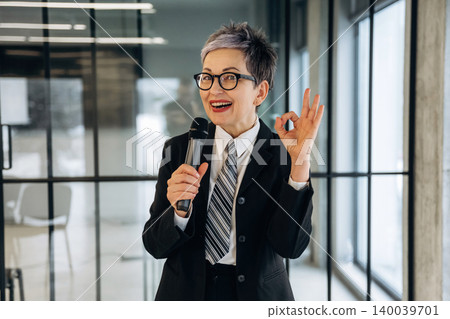 Businesswoman in black suit and glasses confidently speaks into microphone while gesturing with hand inside modern office with large windows and minimalistic design Businesswoman in black suit and glasses confidently speaks into microphone while gesturing with hand inside modern office with large windows and minimalistic design 140039701