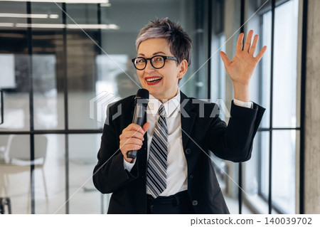 Female business professional with short gray hair wearing a suit and tie holds a microphone while speaking in a modern office setting with glass walls Female business professional with short gray hair wearing a suit and tie holds a microphone while speaking in a modern office setting with glass walls 140039702