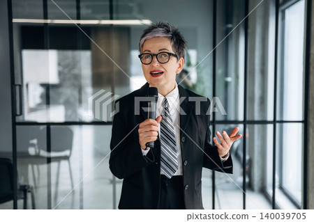 Businesswoman with short gray hair in black suit holding microphone while speaking in modern office with glass walls and contemporary furniture visible in the background Businesswoman with short gray hair in black suit holding microphone while speaking in modern office with glass walls and contemporary furniture visible in the background 140039705