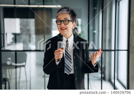 Businesswoman with short gray hair in a black suit and tie holds a microphone while speaking in a modern office with glass walls and contemporary furniture 140039706