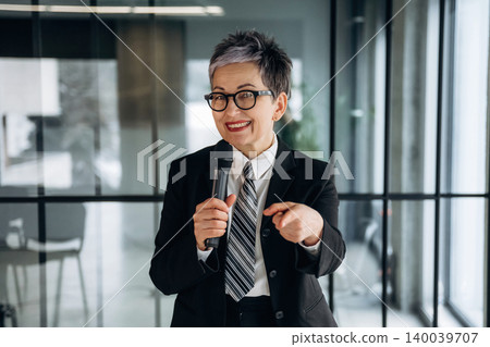 Businesswoman with short gray hair in a black suit holds a microphone while speaking confidently in a modern office setting with glass walls and greenery Businesswoman with short gray hair in a black suit holds a microphone while speaking confidently in a modern office setting with glass walls and greenery 140039707