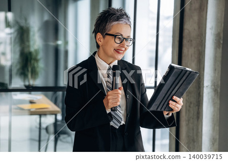 Businesswoman in formal attire holding a tablet and microphone while smiling, standing in a modern office with large windows and a contemporary workspace visible 140039715