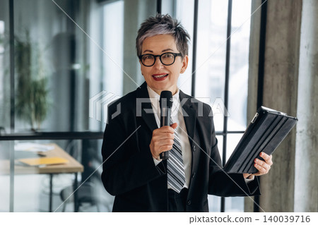 Businesswoman with short gray hair wearing glasses holds a microphone and tablet while standing in a modern office with large windows and a desk in the background Businesswoman with short gray hair wearing glasses holds a microphone and tablet while standing in a modern office with large windows and a desk in the background 140039716