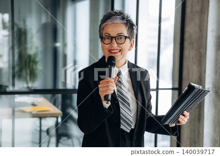 Businesswoman with short gray hair in a black suit holds a microphone and tablet, smiling in a modern office with glass walls and a wooden table in the background Businesswoman with short gray hair in a black suit holds a microphone and tablet, smiling in a modern office with glass walls and a wooden table in the background 140039717