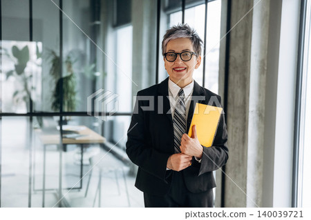 Business professional with short gray hair wearing a black suit and glasses holds a yellow folder while standing in a modern office with large windows and a minimalist design Business professional with short gray hair wearing a black suit and glasses holds a yellow folder while standing in a modern office with large windows and a minimalist design 140039721
