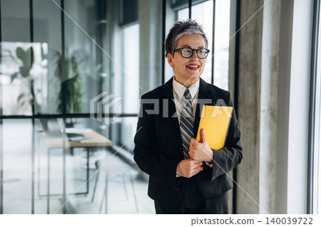 Businesswoman in a black suit holding a yellow folder stands in a modern office with large windows and minimalist furniture, showcasing a professional environment Businesswoman in a black suit holding a yellow folder stands in a modern office with large windows and minimalist furniture, showcasing a professional environment 140039722
