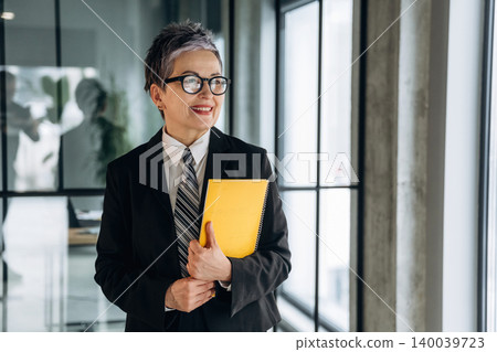 Businesswoman with short gray hair wearing a black suit and glasses holds a yellow notebook while standing in a modern office corridor with large windows 140039723