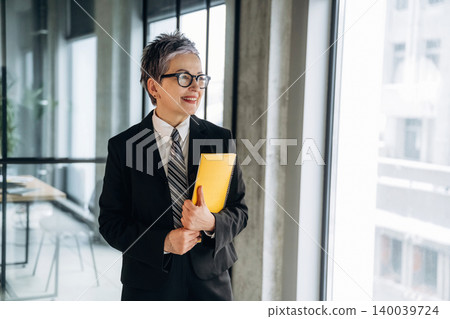 Mature Asian businesswoman in formal suit holding yellow folder stands by large windows in modern office space with glass walls and contemporary furniture Mature Asian businesswoman in formal suit holding yellow folder stands by large windows in modern office space with glass walls and contemporary furniture 140039724