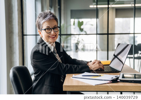 Businesswoman with short gray hair wearing glasses works on laptop at modern office desk with documents and stationery in a bright, spacious environment Businesswoman with short gray hair wearing glasses works on laptop at modern office desk with documents and stationery in a bright, spacious environment 140039729