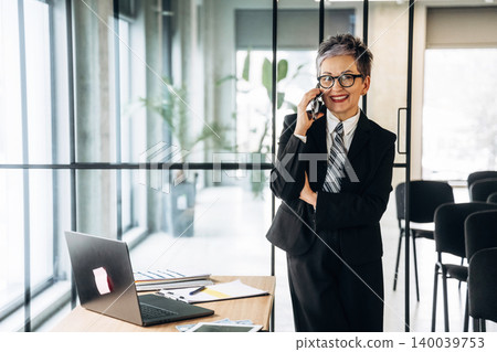Businesswoman in black suit talking on smartphone while standing at desk with laptop and documents in modern office with glass walls and plants 140039753