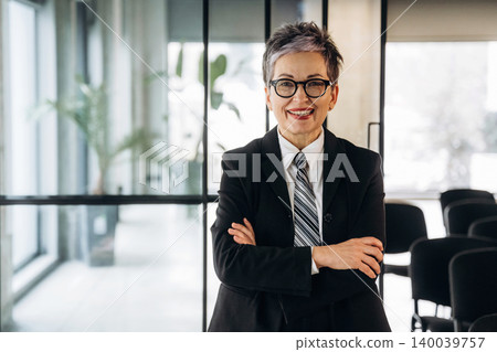 Businesswoman with short gray hair wearing a black suit and glasses stands confidently with arms crossed in a modern office space with glass walls and chairs Businesswoman with short gray hair wearing a black suit and glasses stands confidently with arms crossed in a modern office space with glass walls and chairs 140039757