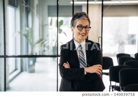 Businesswoman with short gray hair wearing glasses and a black suit stands confidently with arms crossed in a modern office space with glass walls and chairs 140039758