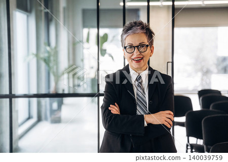 Businesswoman with short gray hair wearing a black suit and tie stands confidently with arms crossed in a modern office space with glass walls and chairs Businesswoman with short gray hair wearing a black suit and tie stands confidently with arms crossed in a modern office space with glass walls and chairs 140039759