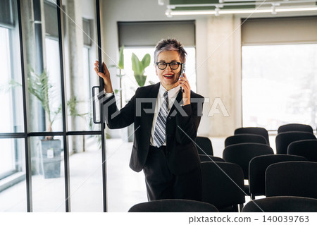 Businesswoman in formal attire smiling while talking on a smartphone, standing by a glass door in a modern office space with chairs and plants visible Businesswoman in formal attire smiling while talking on a smartphone, standing by a glass door in a modern office space with chairs and plants visible 140039763