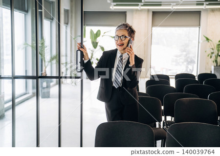 Businesswoman in black suit talking on smartphone while standing by glass door in modern office with empty chairs and indoor plants in the background Businesswoman in black suit talking on smartphone while standing by glass door in modern office with empty chairs and indoor plants in the background 140039764