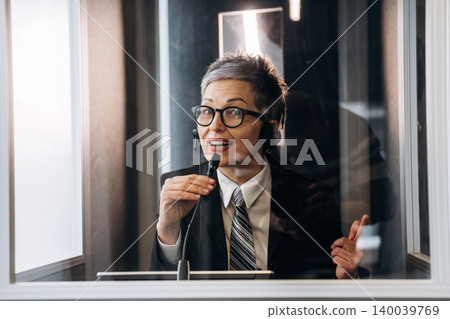 Female interpreter wearing headset speaks into microphone inside soundproof booth, with glass walls and professional office environment visible in the background Female interpreter wearing headset speaks into microphone inside soundproof booth, with glass walls and professional office environment visible in the background 140039769