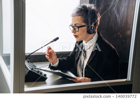 Female interpreter wearing headset speaks into microphone while seated at desk with audio equipment in a soundproof booth during a conference setting Female interpreter wearing headset speaks into microphone while seated at desk with audio equipment in a soundproof booth during a conference setting 140039772