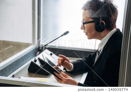 Female interpreter wearing headset and suit uses tablet while working in soundproof booth during conference, microphone and control panel visible in the background Female interpreter wearing headset and suit uses tablet while working in soundproof booth during conference, microphone and control panel visible in the background 140039774