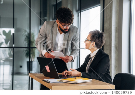 Male and female business professionals discussing project details at a modern office desk with a laptop, documents, and large windows providing natural light Male and female business professionals discussing project details at a modern office desk with a laptop, documents, and large windows providing natural light 140039789