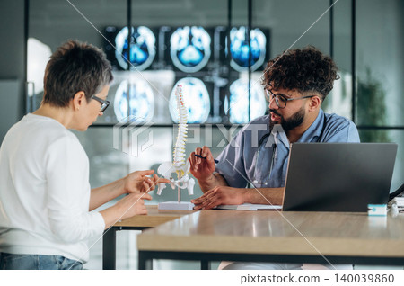 Male healthcare professional explains spinal anatomy to female patient using anatomical model in modern medical office with medical imaging in background Male healthcare professional explains spinal anatomy to female patient using anatomical model in modern medical office with medical imaging in background 140039860