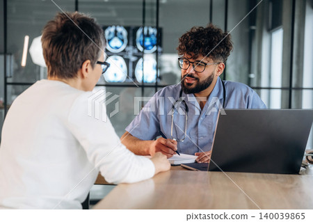 Male healthcare professional in blue scrubs discusses medical results with a female patient at a desk, laptop and medical images visible in the background Male healthcare professional in blue scrubs discusses medical results with a female patient at a desk, laptop and medical images visible in the background 140039865